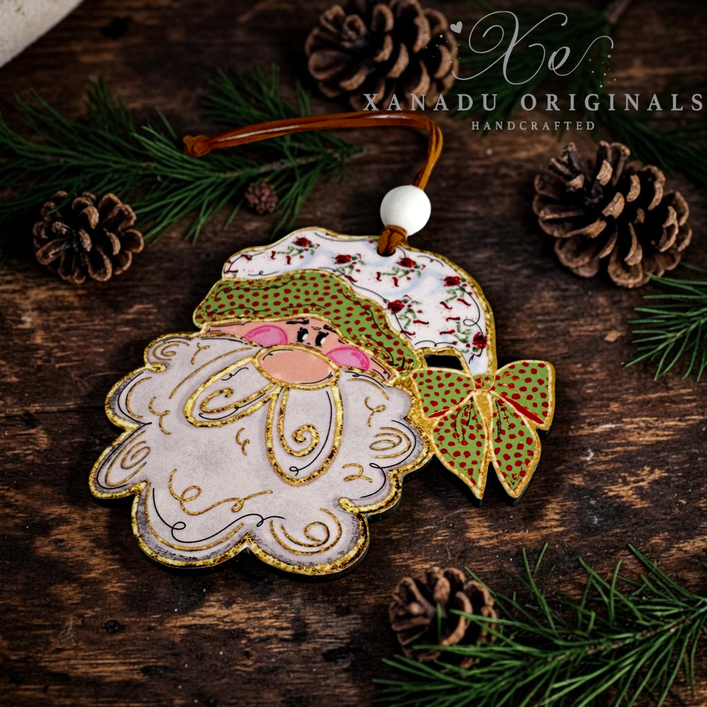 A handmade wooden Christmas ornament shaped like Santa's face and hat, hanging from a brown cord with a white bead. The ornament features gold embroidery, green patterned details, and pink accents, displayed on a rustic dark wood background with pine needles and pinecones.