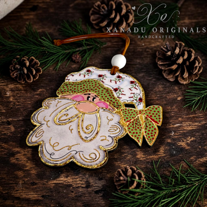 A handmade wooden Christmas ornament shaped like Santa's face and hat, hanging from a brown cord with a white bead. The ornament features gold embroidery, green patterned details, and pink accents, displayed on a rustic dark wood background with pine needles and pinecones.