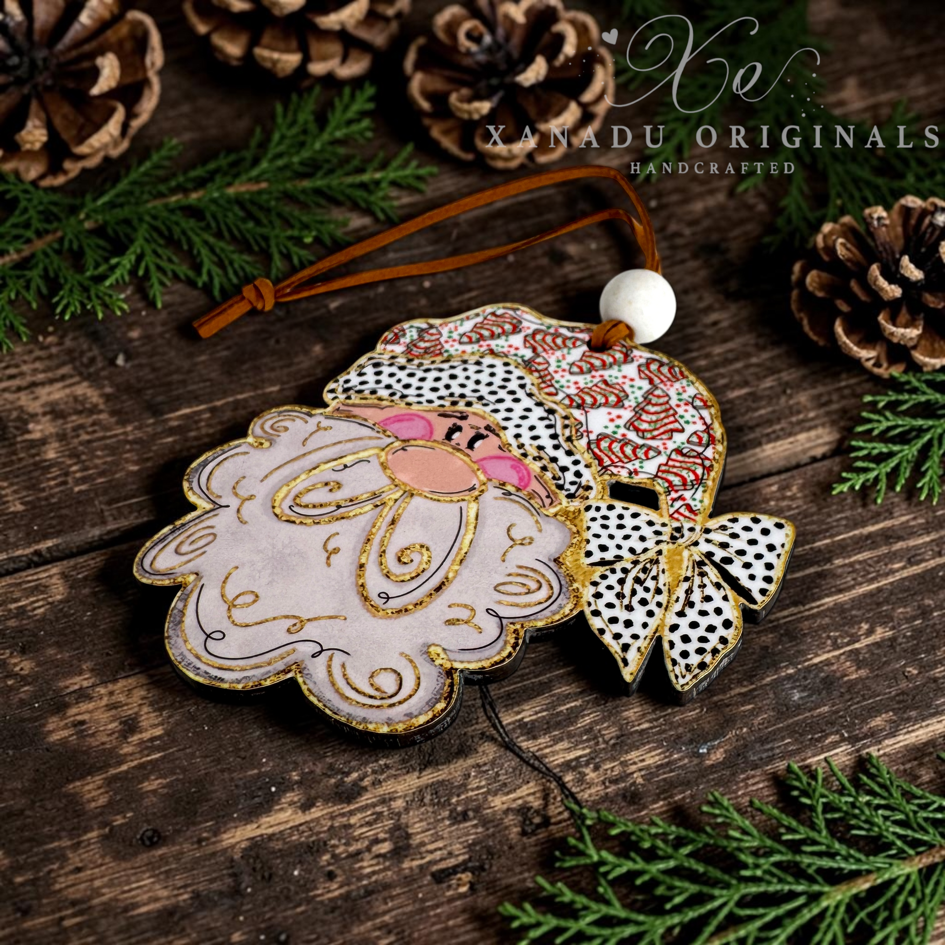 A handcrafted Christmas ornament featuring a stylized Santa face with a pink beard, gold glitter outlines, and a Santa hat with black-and-white polka dots and red accents. The ornament hangs from a brown leather string with a white bead, photographed on a rustic dark wood surface surrounded by pine branches and pinecones.
