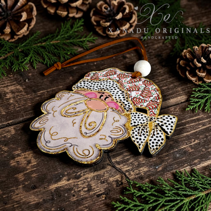 A handcrafted Christmas ornament featuring a stylized Santa face with a pink beard, gold glitter outlines, and a Santa hat with black-and-white polka dots and red accents. The ornament hangs from a brown leather string with a white bead, photographed on a rustic dark wood surface surrounded by pine branches and pinecones.