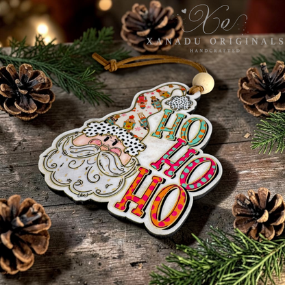 A handcrafted wooden Christmas ornament shaped like Santa's face with 'HO HO HO' lettering, displayed on a rustic wooden surface surrounded by pine branches, pinecones, and warm holiday lights. The ornament features a white base with colorful patterned details and a brown hanging cord.