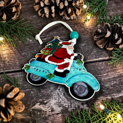 A Christmas ornament shaped like a turquoise vintage car with Santa Claus riding on top while carrying a decorated tree. The ornament is photographed on a rustic wooden surface surrounded by pine branches, pinecones, and warm string lights.