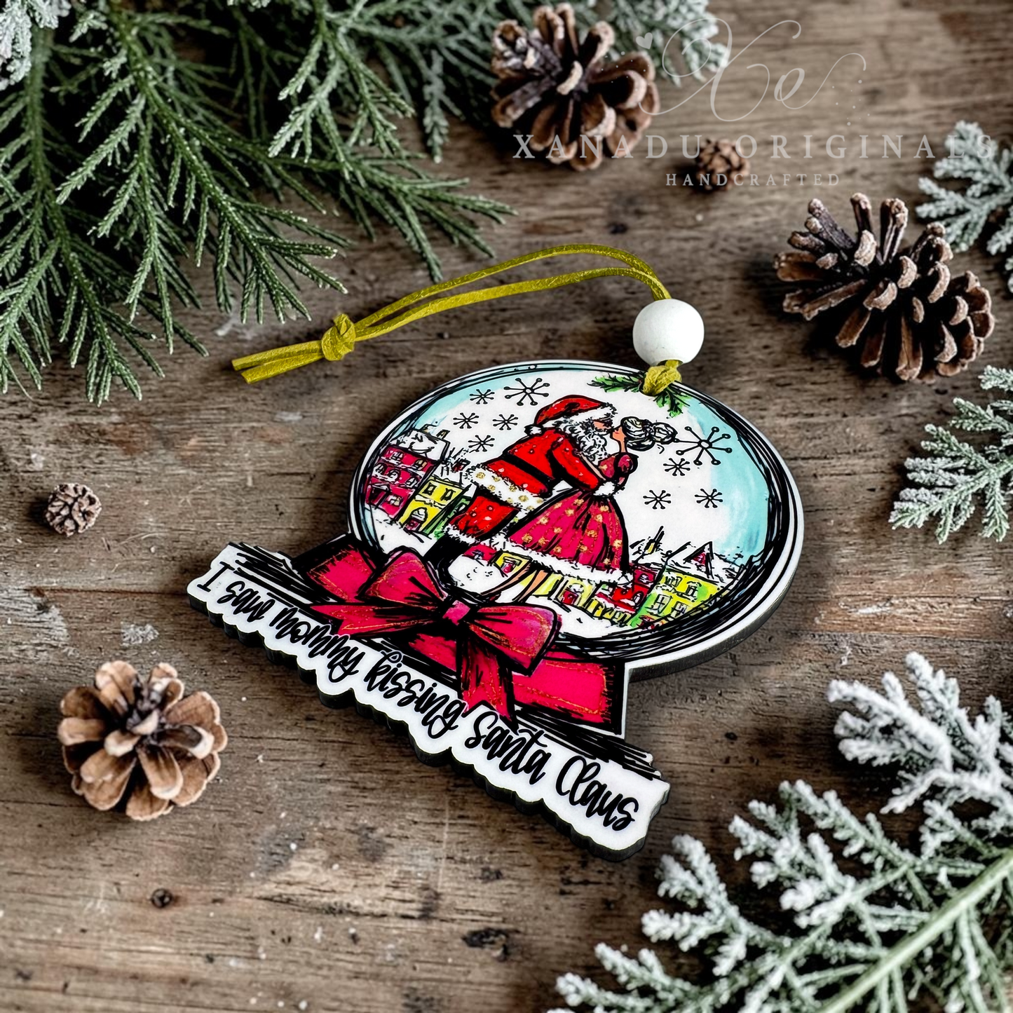 A Christmas ornament featuring an illustration of Santa Claus in a snowy village scene, with a red bow and the text 'I saw mommy kissing Santa Claus' on a rustic wooden background with pine branches and pinecones.