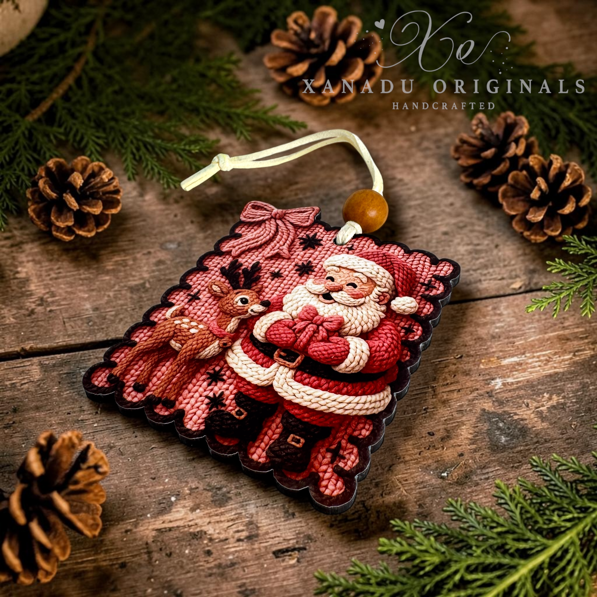 A handcrafted Christmas ornament featuring Santa Claus and a reindeer, styled on a rustic wooden surface with pine branches, pinecones, and a brand watermark for Xanadu Originals.
