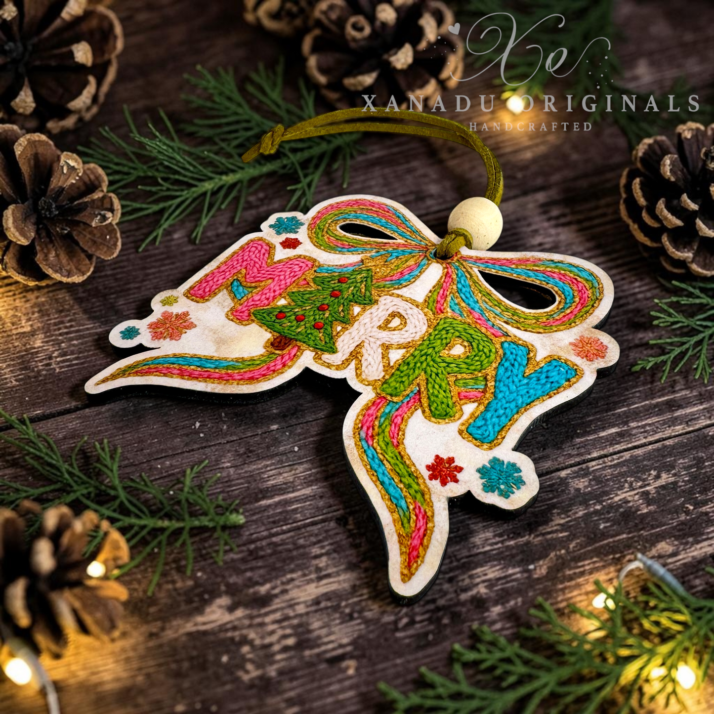 A handcrafted wooden Christmas ornament with colorful embroidered 'MERRY' lettering and a bow design, hanging on a rustic dark wood surface with pine branches, pinecones, and warm string lights. The brand name 'XANADU ORIGINALS' is watermarked in the upper right.