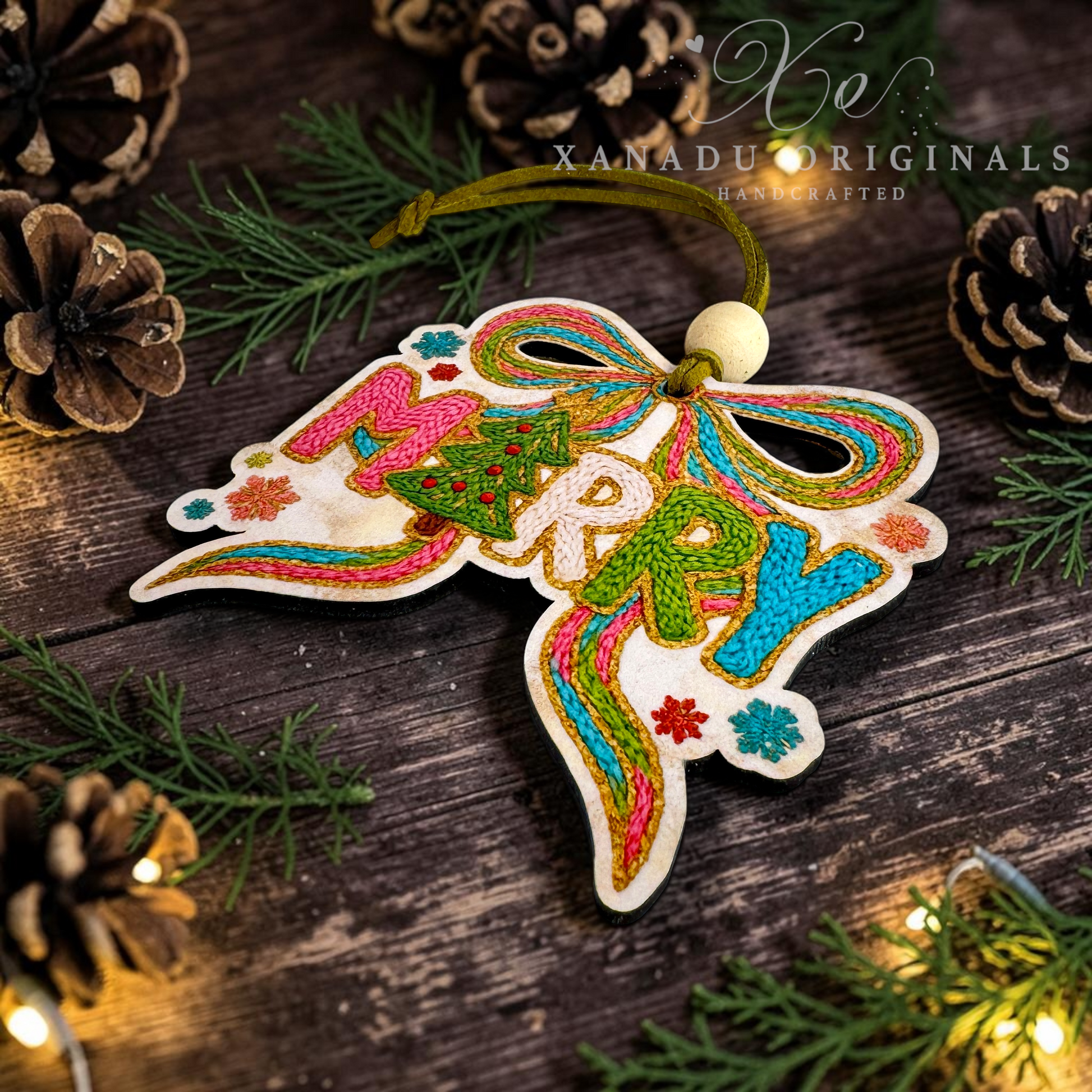 A handcrafted wooden Christmas ornament with colorful embroidered 'MERRY' lettering and a bow design, hanging on a rustic dark wood surface with pine branches, pinecones, and warm string lights. The brand name 'XANADU ORIGINALS' is watermarked in the upper right.