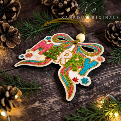 A handcrafted wooden Christmas ornament with colorful embroidered 'MERRY' lettering and a bow design, hanging on a rustic dark wood surface with pine branches, pinecones, and warm string lights. The brand name 'XANADU ORIGINALS' is watermarked in the upper right.