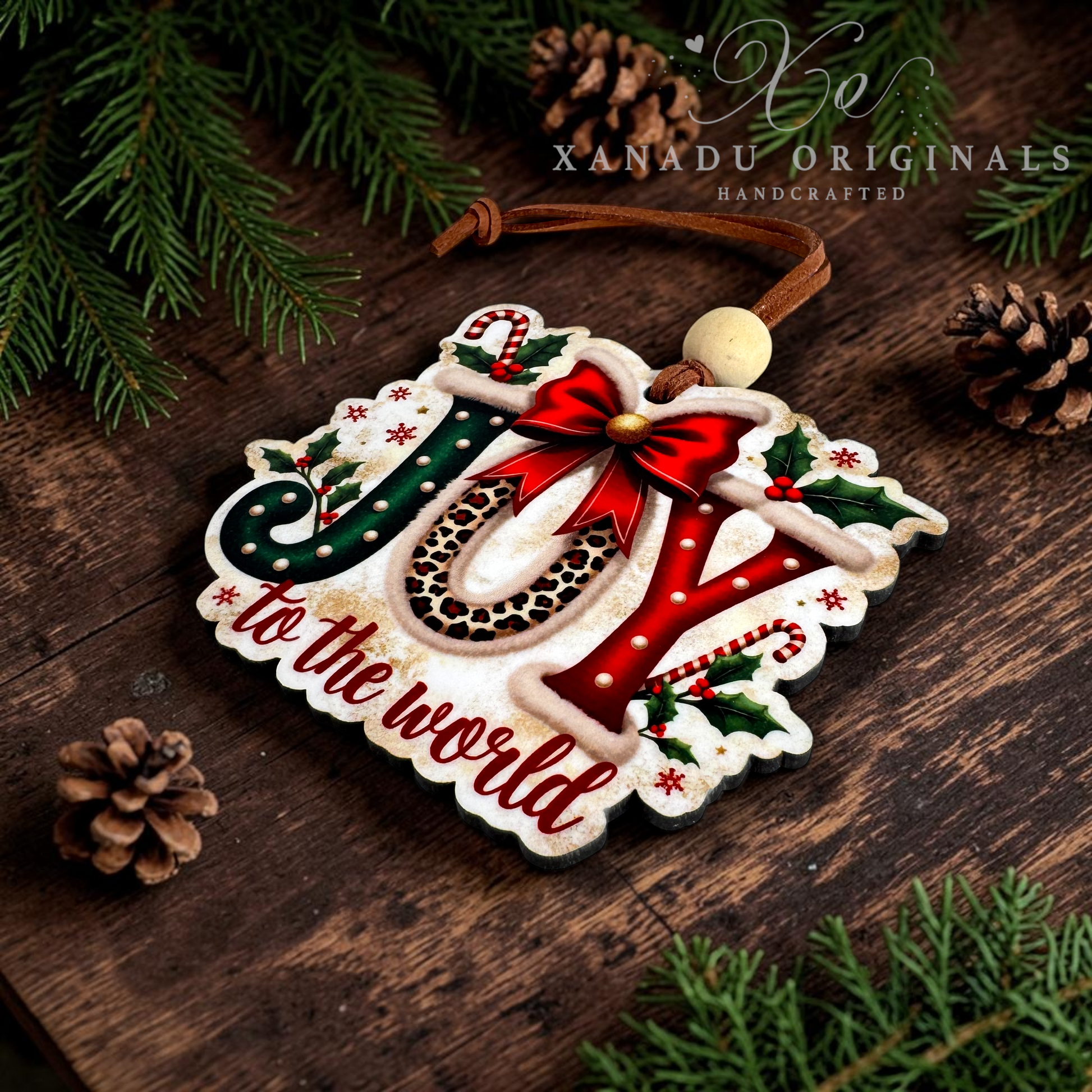 A handcrafted wooden Christmas ornament spelling 'JOY' with 'to the world' below, decorated with red, green, and leopard-print details, hanging on a rustic dark wood surface with pine branches and pinecones. A watermark reads 'XANADU ORIGINALS HANDCRAFTED'.