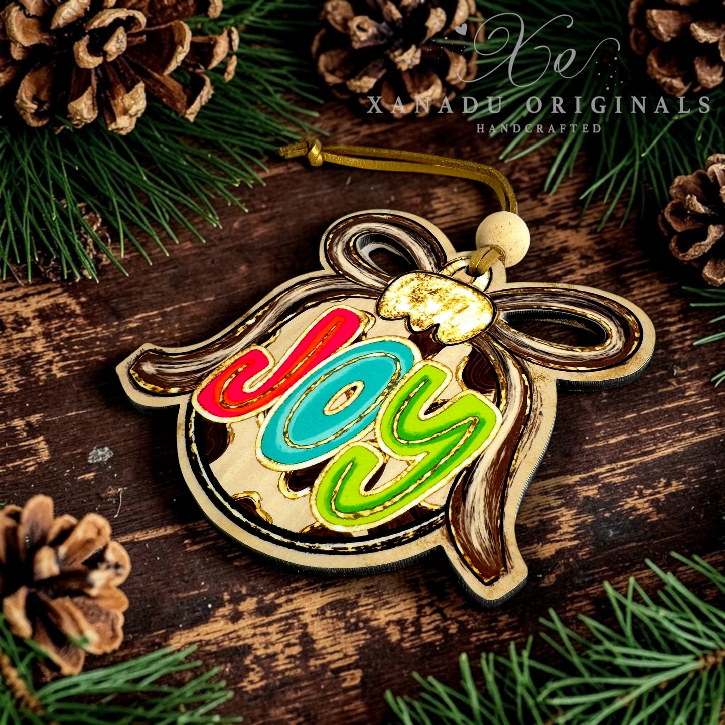 A wooden Christmas ornament shaped like a bauble with the word 'joy' in colorful red, blue, and green letters, displayed on a rustic dark wood surface with pine branches and pinecones. A watermark reads 'XANADU ORIGINALS HANDCRAFTED'.