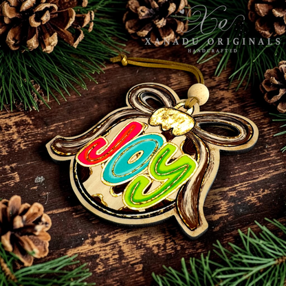 A wooden Christmas ornament shaped like a bauble with the word 'joy' in colorful red, blue, and green letters, displayed on a rustic dark wood surface with pine branches and pinecones. A watermark reads 'XANADU ORIGINALS HANDCRAFTED'.