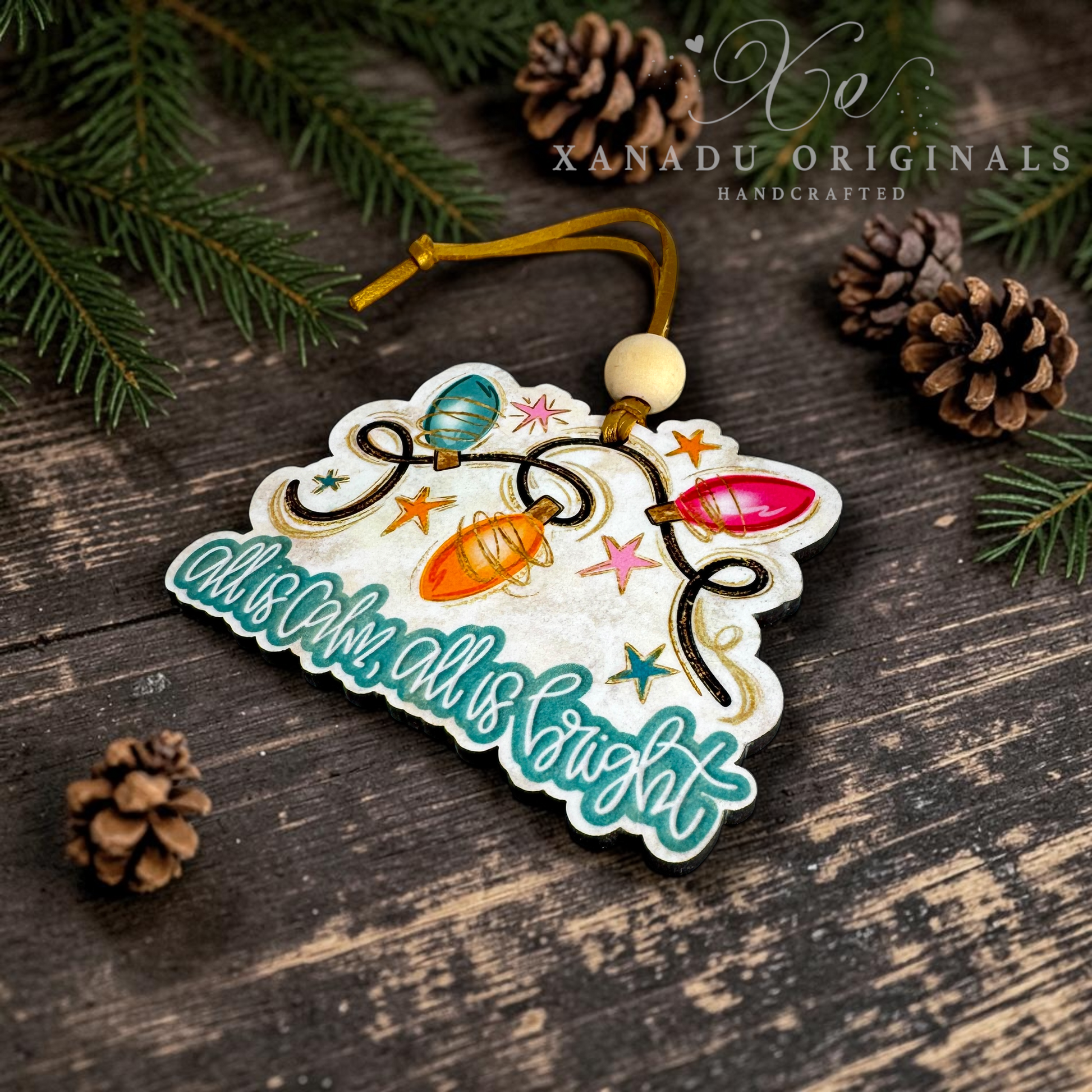 A decorative Christmas ornament shaped like a tree branch with colorful hanging lights and the phrase “advent all is bright,” photographed on a dark wood surface with pine branches and pinecones. A watermark reading XANADU ORIGINALS HANDCRAFTED appears in the upper right.