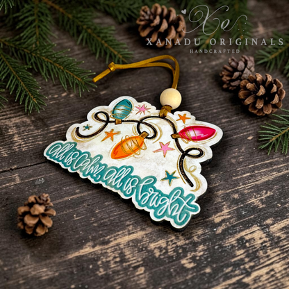 A decorative Christmas ornament shaped like a tree branch with colorful hanging lights and the phrase “advent all is bright,” photographed on a dark wood surface with pine branches and pinecones. A watermark reading XANADU ORIGINALS HANDCRAFTED appears in the upper right.