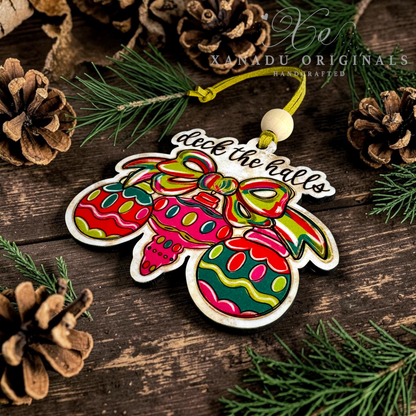 A colorful Christmas ornament shaped like three hanging ornaments with the phrase “dick the hells” in script, styled on a rustic wooden surface with pine branches and pinecones. A watermark reading “XANADU ORIGINAL HANDCRAFTED” appears near the top.
