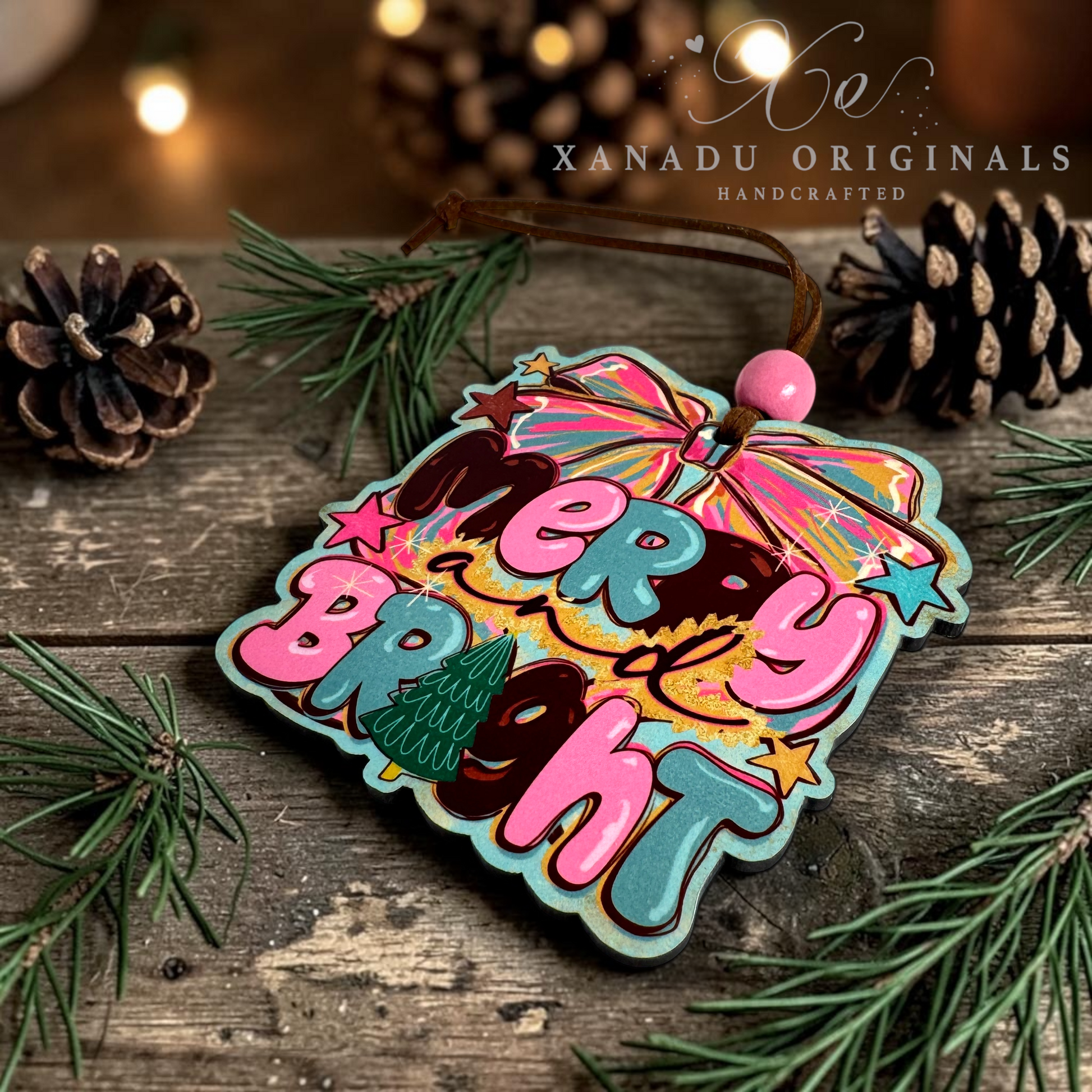 A colorful Christmas ornament with the words “Merry Bright” in pink and teal lettering, decorated with a rainbow umbrella, stars, and a small Christmas tree, resting on a rustic wooden surface with pine needles and pinecones. The brand name “XANADU ORIGINALS” is visible in the upper right.