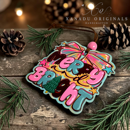 A colorful Christmas ornament with the words “Merry Bright” in pink and teal lettering, decorated with a rainbow umbrella, stars, and a small Christmas tree, resting on a rustic wooden surface with pine needles and pinecones. The brand name “XANADU ORIGINALS” is visible in the upper right.