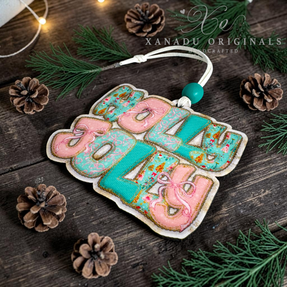 A decorative hanging ornament spelling 'Holly Jolly' in pink and turquoise lettering with a white cord and turquoise bead, styled on a dark wood surface with pine branches, pinecones, and fairy lights. A watermark reads 'XANADU ORIGINALS HANDCRAFTED'.