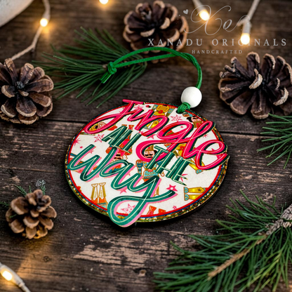 A round wooden Christmas ornament with the phrase 'Jingle All the Way' in colorful script, hanging from a green cord with a white bead. The ornament is styled on a rustic dark wood surface with pine branches, pinecones, and warm string lights.