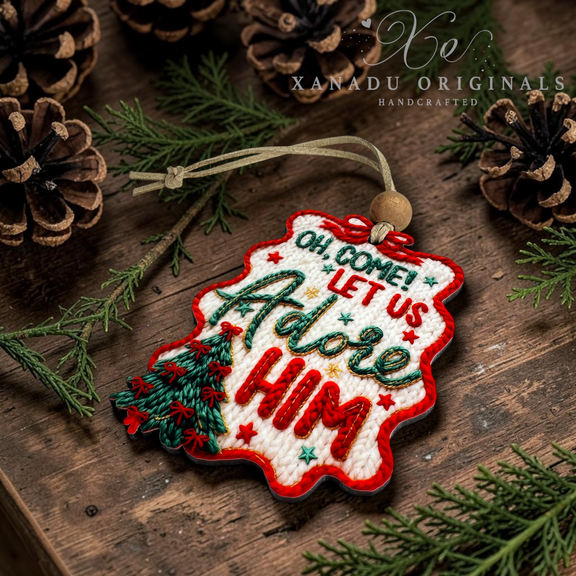 An embroidered Christmas ornament shaped like a tree, featuring the phrase 'Oh Come! Let Us Adore Him' in red and green thread, resting on a rustic wooden surface surrounded by pine branches and pinecones. The brand name 'XANADU ORIGINALS' is visible in the top right corner.