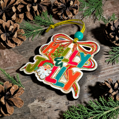 A wooden Christmas ornament shaped like a bow and ribbon, featuring the words 'Holly & Jolly' in colorful lettering and a small Santa illustration, displayed on a rustic wooden surface with pinecones and evergreen branches.
