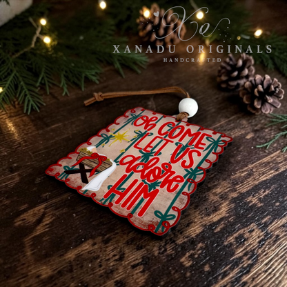 A handcrafted wooden Christmas ornament with a scalloped edge, featuring the words 'oh Come Let Us adore Him' in red and green lettering, It hangs from a brown string with a white bead.