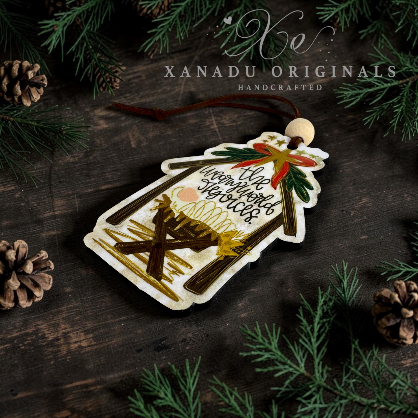 A handcrafted wooden Nativity scene ornament with a hanging loop, featuring a gold angel, green wreath, and the words 'The weary world rejoices' on a dark rustic wooden background with pine branches and pinecones. 'XANADU ORIGINALS HANDCRAFTED' is watermarked at the top.