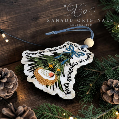 Handcrafted Christmas ornament shaped like a pine tree with a nativity scene and personalized text, displayed on a rustic wooden surface with pine branches, pinecones, and warm string lights. The brand name 'XANADU ORIGINALS' is visible in the upper right.