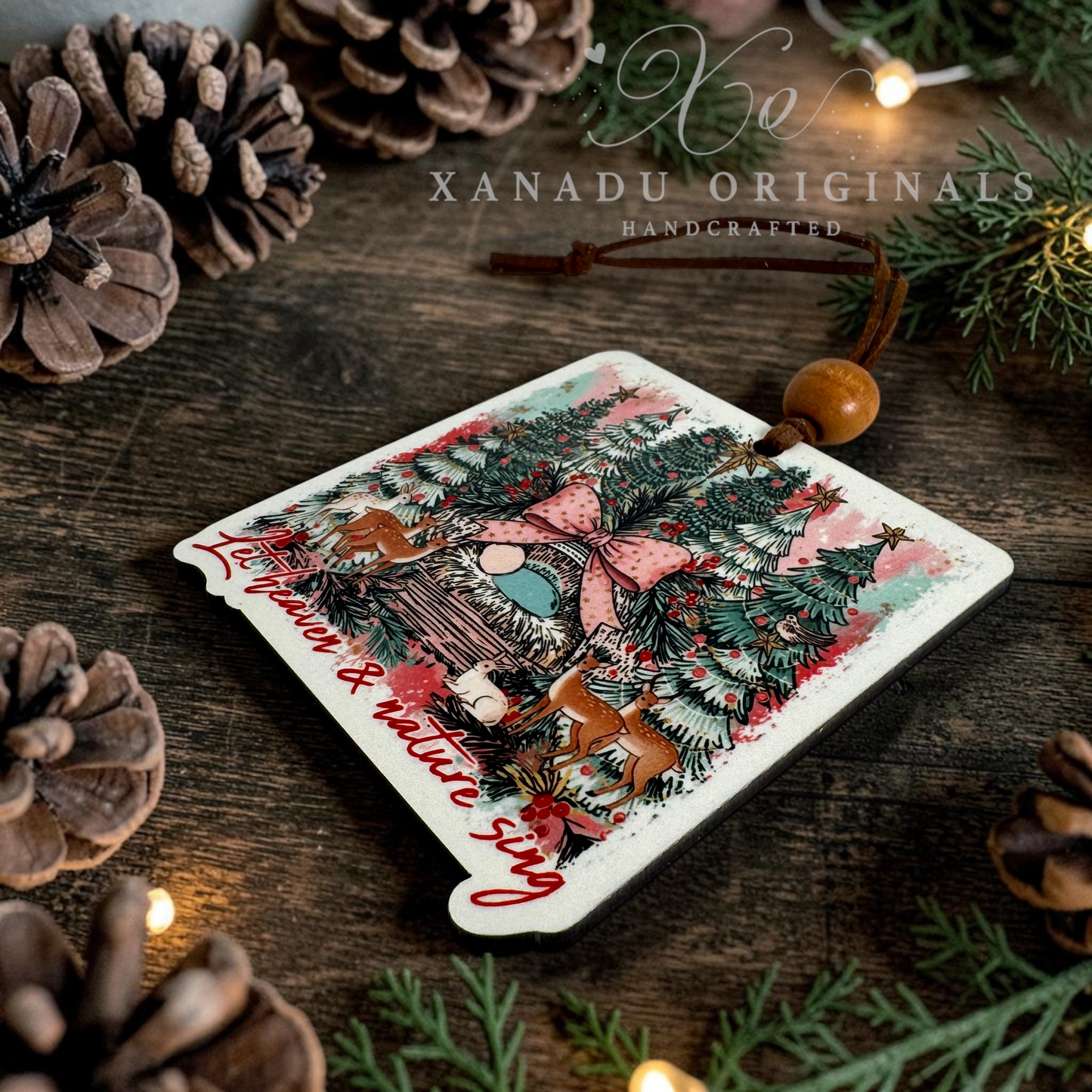 A square Christmas ornament with rounded corners, featuring a festive illustration of a cozy cabin, pine trees, and deer, with the text 'Let Heaven & Nature Sing' in red script. The ornament is decorated with a wooden bead and hanging string, resting on a rustic wooden surface surrounded by pinecones and evergreen branches.