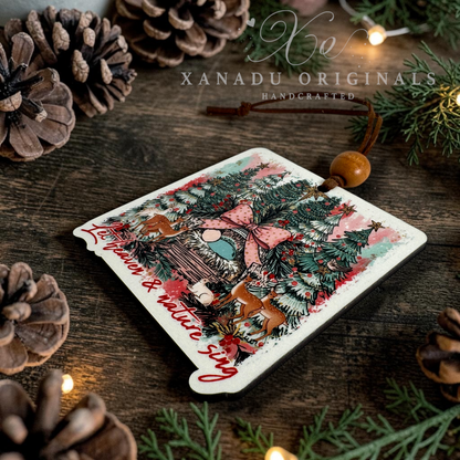 A square Christmas ornament with rounded corners, featuring a festive illustration of a cozy cabin, pine trees, and deer, with the text 'Let Heaven & Nature Sing' in red script. The ornament is decorated with a wooden bead and hanging string, resting on a rustic wooden surface surrounded by pinecones and evergreen branches.