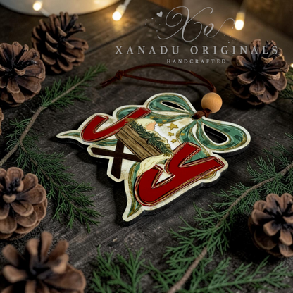 A handcrafted wooden Christmas ornament with the word Joy with the O being baby Jesus decorated with a miniature Christmas tree and pinecones, displayed on a dark rustic wooden surface with pine branches and pinecones.