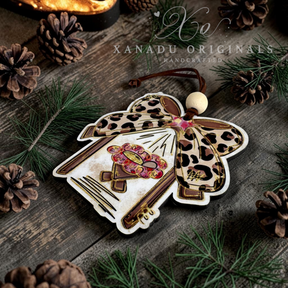 A handcrafted Christmas ornament shaped like a manger with leopard-print bow, a wooden bead head, and a colorful embroidered bodice, displayed on a rustic wooden surface with pine needles and pinecones. The brand name 'XANADU ORIGINALS' is watermarked across the top.