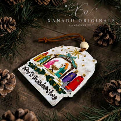 A handcrafted Christmas ornament featuring a nativity scene with colorful figures, a star, and the text 'Glory to the Newborn King,' displayed on a rustic wooden surface with pine branches and pinecones. The brand name 'XANADU ORIGINALS' is visible at the top.