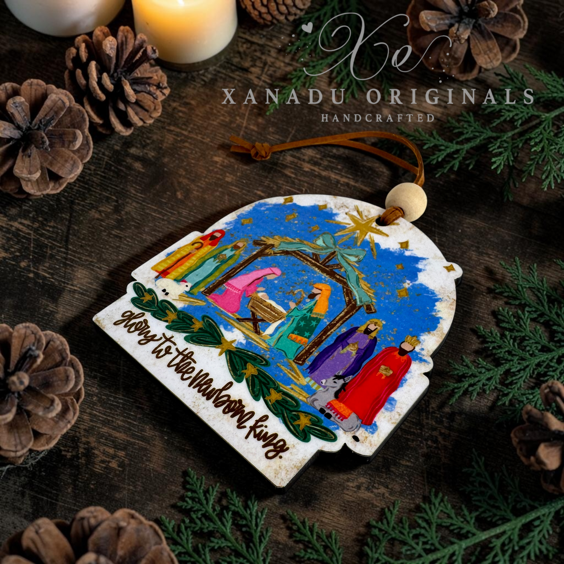 A handcrafted Christmas ornament featuring a colorful nativity scene with the text 'Glory to the Newborn King' on a distressed white background, styled on a dark wooden surface with pinecones, candles, and greenery. The brand name 'XANADU ORIGINALS' is visible at the top.