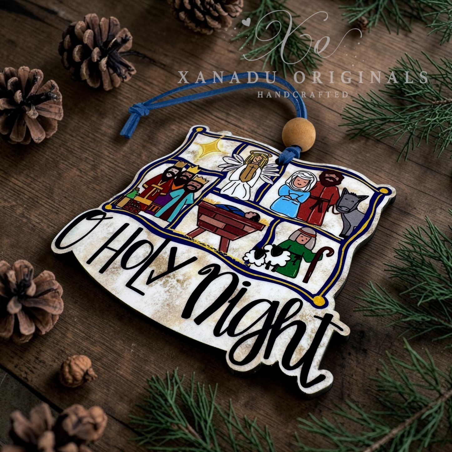 A decorative Christmas ornament shaped like a rustic wooden sign with the words “O Holy Night” in black script, featuring illustrated scenes from the nativity on a weathered wood background. The ornament hangs from a blue string with a round wooden bead, styled on a dark wooden surface with pine branches, pinecones, and evergreen needles.