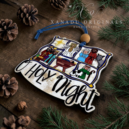 A decorative Christmas ornament shaped like a rustic wooden sign with the words “O Holy Night” in black script, featuring illustrated scenes from the nativity on a weathered wood background. The ornament hangs from a blue string with a round wooden bead, styled on a dark wooden surface with pine branches, pinecones, and evergreen needles.
