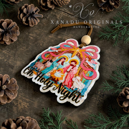A handcrafted Christmas ornament shaped like a stable with a colorful nativity scene and the words 'Oh Holy Night' in gold glitter script. The ornament is displayed on a dark wooden surface surrounded by pine needles, pinecones, and a wooden bead hanging loop.