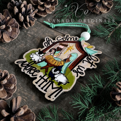 A handcrafted wooden Christmas ornament featuring a nativity scene with a baby in a manger, sheep, and the words 'Oh come let us adore HIM!' on a rustic dark wood background with pine needles and pinecones.
