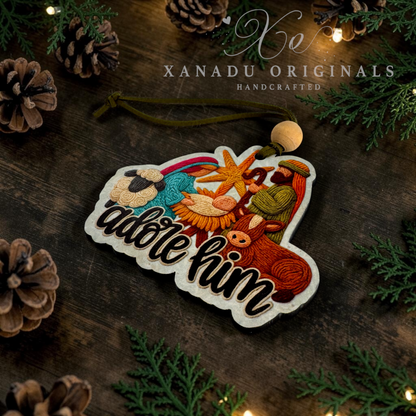 A handcrafted wooden Nativity ornament with embroidered figures and the words 'adore him,' displayed on a dark rustic wooden surface with pinecones, greenery, and warm string lights. The brand name 'XANADU ORIGINALS' is visible at the top.