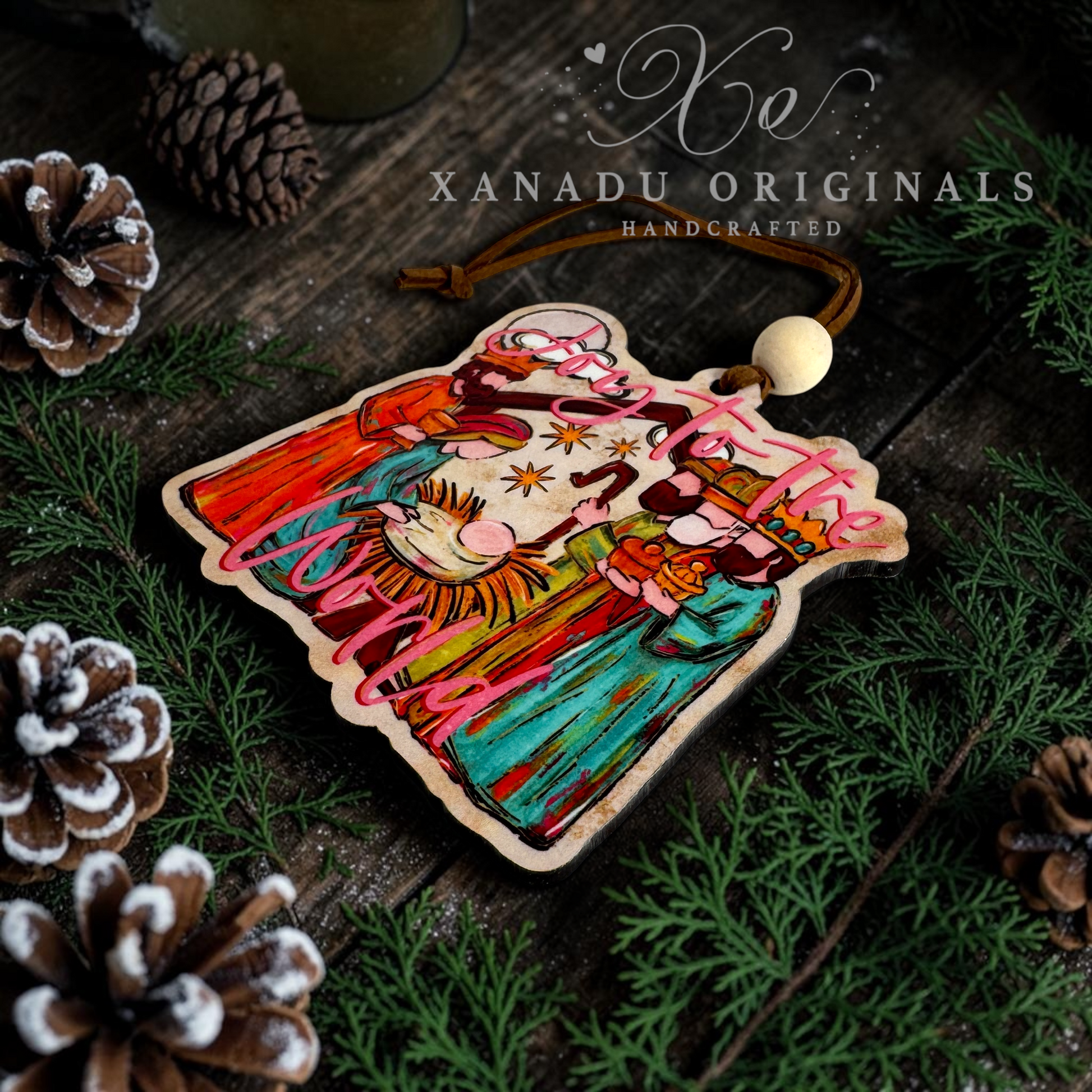 A handcrafted wooden Christmas ornament featuring a colorful illustration of a manger scene, with the text 'Joy to the World' and 'XANADU ORIGINALS HANDCRAFTED' printed on it. The ornament is displayed on a dark rustic wooden surface surrounded by pine needles, pinecones, and a green mug.