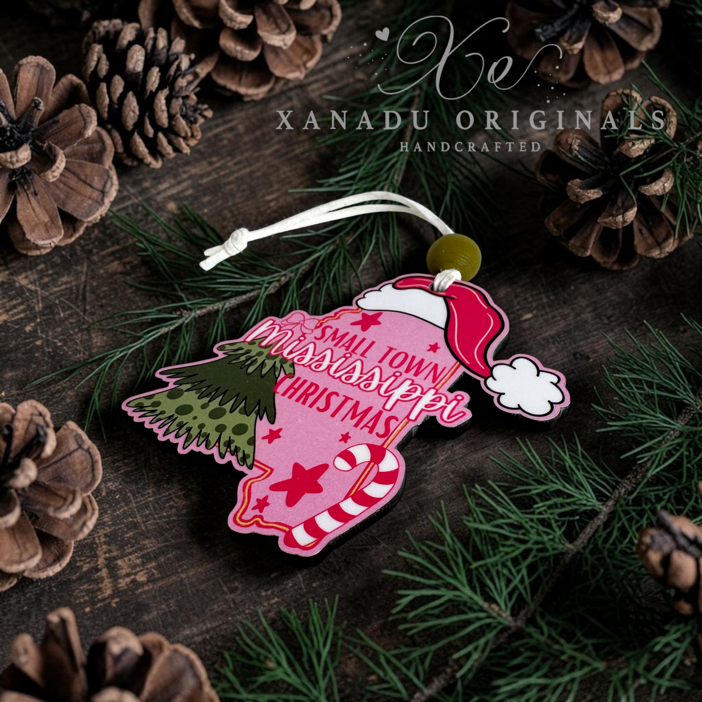A festive Christmas ornament shaped like a bell, featuring a pink body with the words 'Small Town Mississippi Christmas' in red script, a red Santa hat, and a green Christmas tree illustration. The ornament is displayed on a dark wooden surface surrounded by pine needles, pinecones, and a white Xanadu Originals watermark.