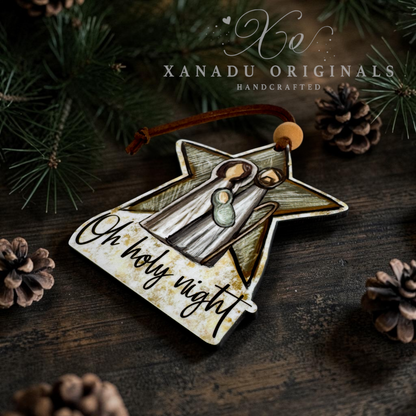 A star-shaped Christmas ornament with a rustic distressed finish, featuring an illustration of an angel and the text 'Oh holy night'. The ornament is displayed on a dark wooden surface surrounded by pine needles and pinecones, with the brand name 'Xanadu Originals' visible at the top.