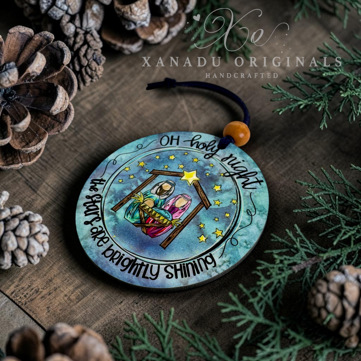 A round wooden Christmas ornament featuring a hand-painted nativity scene with the text 'Oh holy night' and 'The Star is Brightly Shining,' displayed on a rustic wooden surface surrounded by pine needles, pinecones, and a small orange bead hanging from a black cord. Xanadu Originals branding is visible at the top.