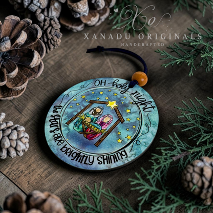 A round wooden Christmas ornament featuring a hand-painted nativity scene with the text 'Oh holy night' and 'The Star is Brightly Shining,' displayed on a rustic wooden surface surrounded by pine needles, pinecones, and a small orange bead hanging from a black cord. Xanadu Originals branding is visible at the top.