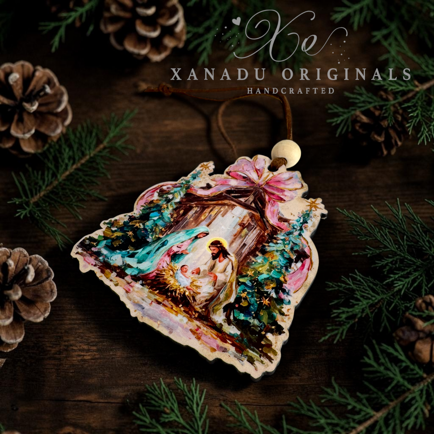 A handcrafted Christmas ornament, featuring a painted nativity scene with Mary, Joseph, baby Jesus, and a pink bow. The ornament is laying on a dark wooden surface with pine branches and pinecones, and the text 'XANADU ORIGINALS HANDCRAFTED' is visible at the top.