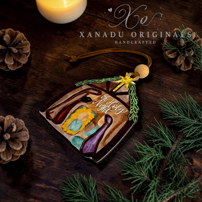A handcrafted wooden Christmas ornament shaped like a house, featuring a nativity scene with the words 'Oh Holy Night' written on it. The ornament is styled on a dark wooden surface with pine branches, pinecones, and a lit candle, with the brand name 'XANADU ORIGINALS' visible at the top.