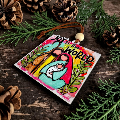 A square wooden ornament featuring a colorful hand-painted manger scene and the text 'Joy to the World,' hanging from a brown cord with a wooden bead. The ornament is styled on a rustic dark wood surface with pine branches and pinecones, and a watermark reading 'XANADU ORIGINALS HANDCRAFTED' appears at the top.