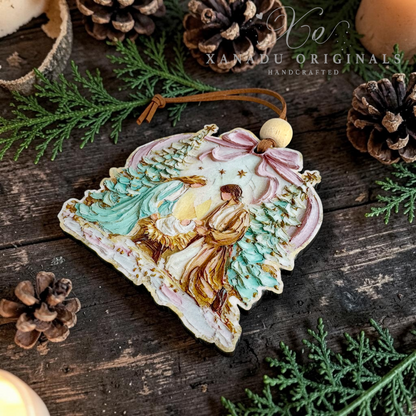 A handcrafted Christmas ornament featuring an angel and baby Jesus in a snowy landscape, styled on a rustic dark wood surface with pinecones, evergreen branches, and a warm candle glow. The ornament has a pink bow, a cream-colored top bead, and a brown hanging string.