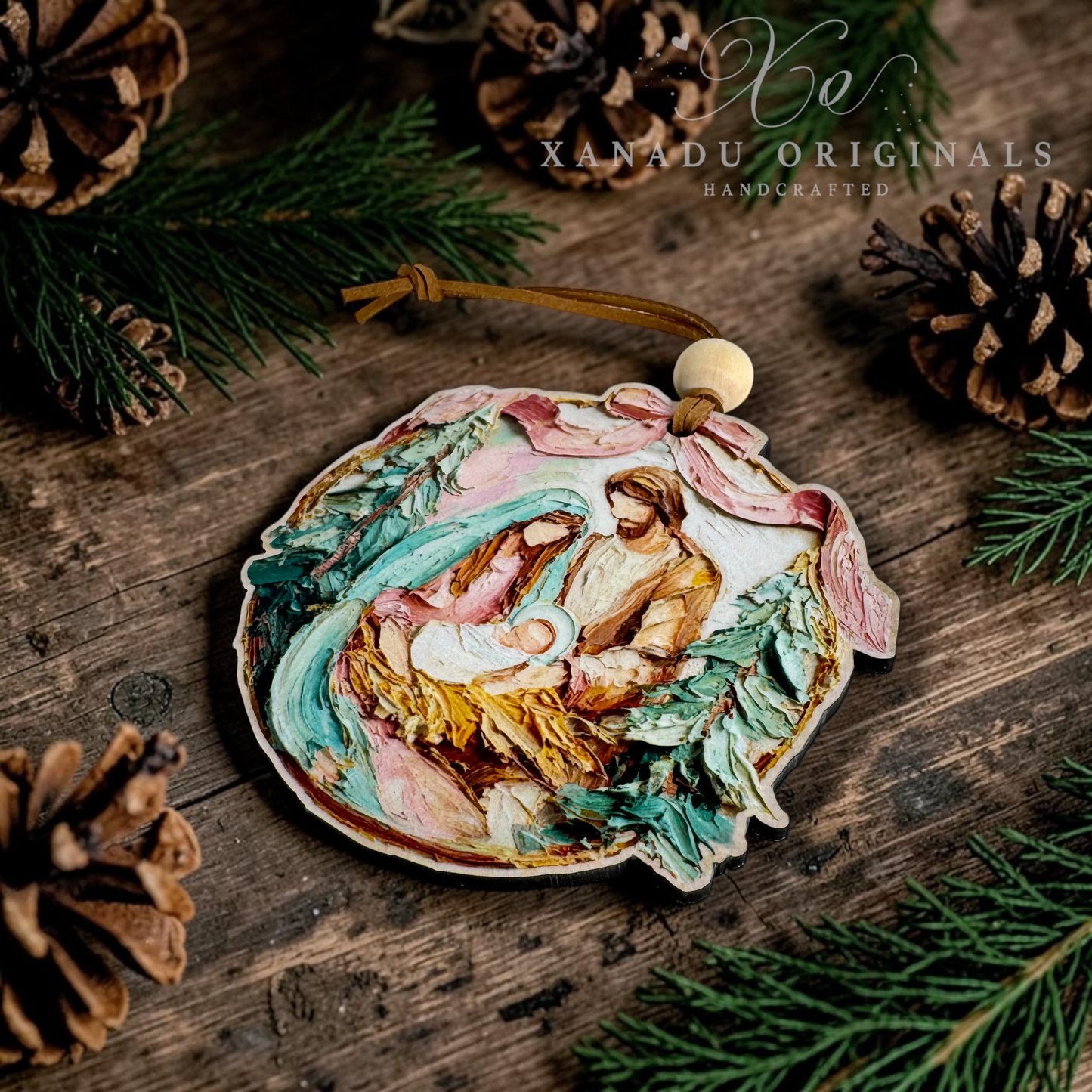 A handcrafted wooden Christmas ornament featuring a painted nativity scene with Mary, Joseph, and baby Jesus, hanging from a brown cord with a wooden bead. The ornament is styled on a rustic wooden surface with pine needles and pinecones, with the brand name 'XANADU ORIGINALS' visible in the upper right.