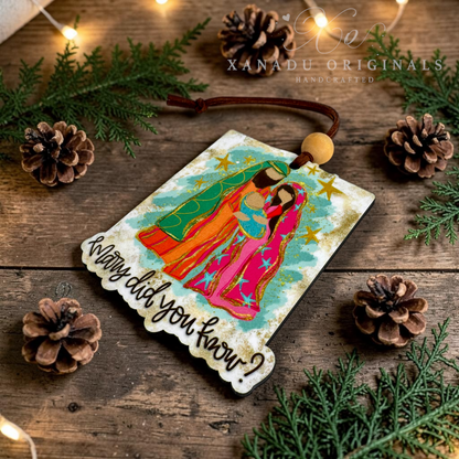 A rectangular Christmas ornament with a scalloped white border, featuring a colorful illustration of Mary and Joseph with the text 'Mary did you know?' and a brown hanging cord, styled on a rustic wooden surface with pinecones, evergreen branches, and warm string lights. A watermark for 'XANADU ORIGINALS HANDCRAFTED' is visible at the top.
