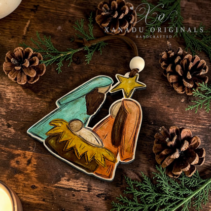 A handcrafted wooden Nativity scene ornament featuring Mary, Joseph, baby Jesus in a manger, and a gold star, displayed on a rustic wooden surface with pinecones and evergreen branches. The brand name 'XANADU ORIGINALS' is visible as a watermark in the upper right.