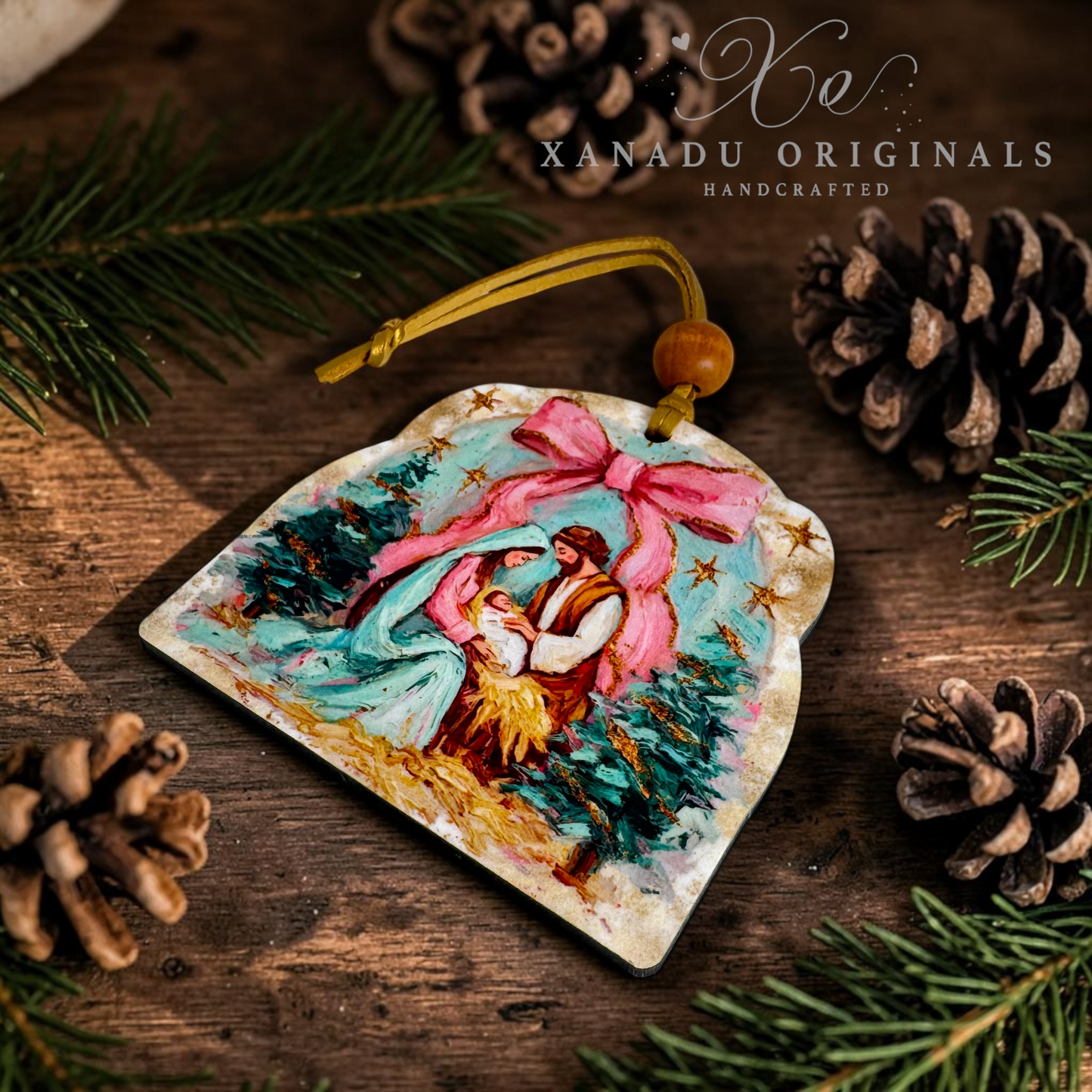 A handcrafted wooden Christmas ornament featuring an illustrated nativity scene with a pink bow, resting on a rustic wooden surface surrounded by pine needles and pinecones. The brand name 'XANADU ORIGINALS' is visible in the upper right corner.